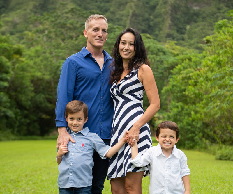 A man a woman stand holding hands with their two toddler sons in a grassy field in front of a mountain.