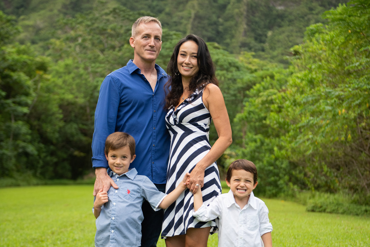 A man a woman stand holding hands with their two toddler sons in a grassy field in front of a mountain.