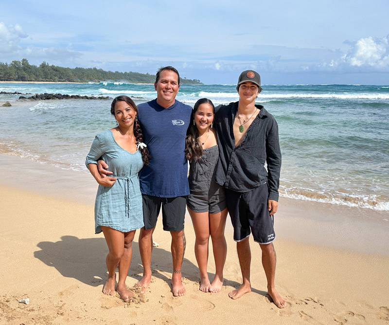 A woman and man stand with their teenage daughter and teenage son on the shore of Hanalei Bay on Kauai.