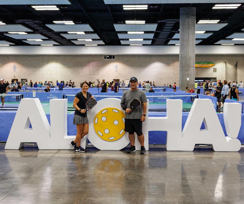 two pickleball players stand in front of a giant ALOHA sign