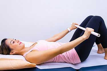 Woman laying on back doing exercise with arms holding a baton at her bent knees..