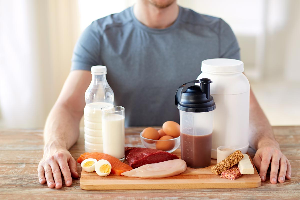 a man sits in front of a wooden tray stacked with milk, chicken, fish, steak, eggs and protein supplements