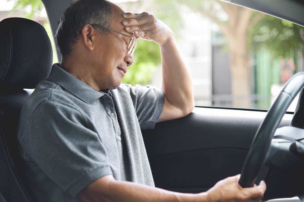 older man holds head while sitting in the driver's seat of a car