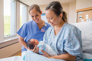Woman in hospital holding newborn baby as a nurse looks on.