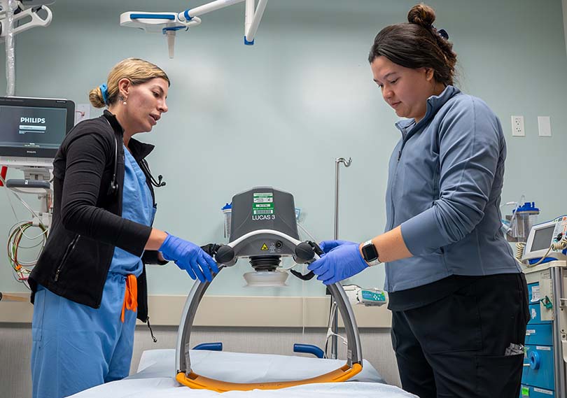 Two medical professionals holding an automated CPR machine in a medical exam room.