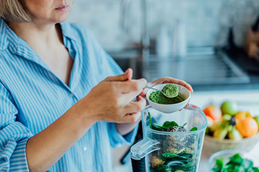 Woman scooping greens powders into a blender full of fresh fruits and vegetables.