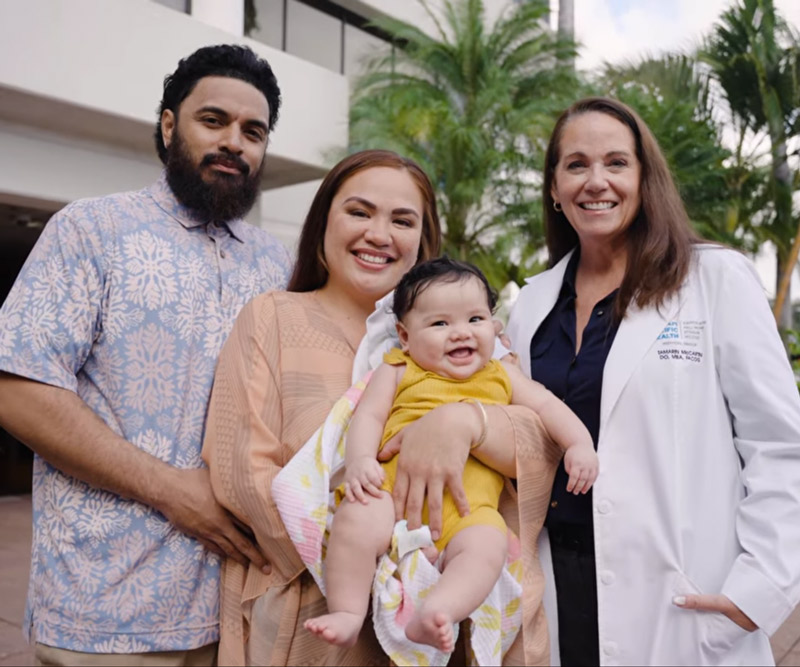 a dad and mom hold their baby girl and stand with a doctor smiling in the courtyard at Pali Momi Medical Center