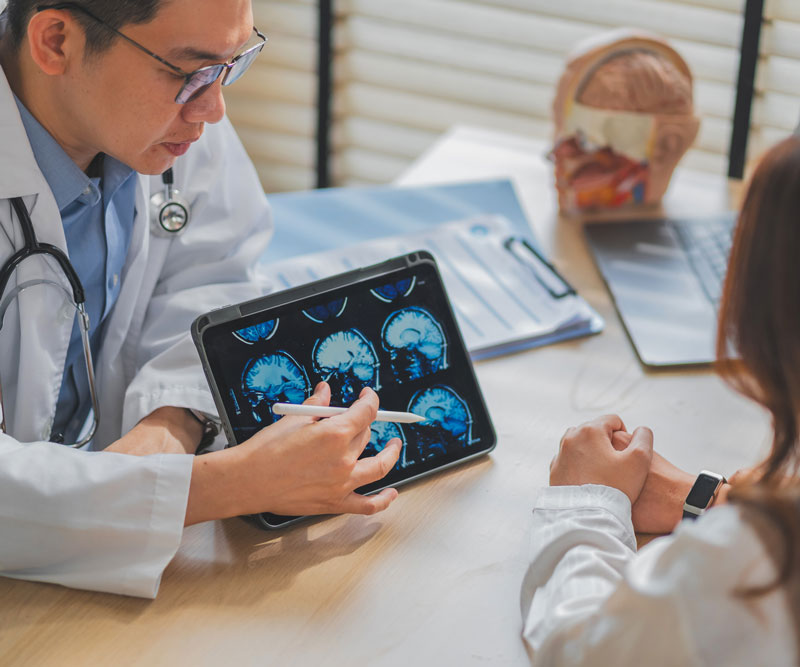 a neurologist sits behind his desk in his office and shows a patient a brain scan on an iPad