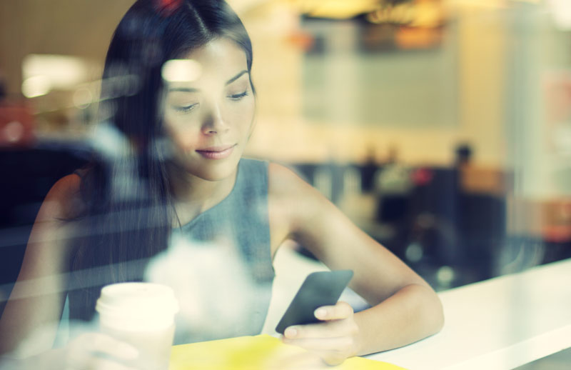 a lady with a cup of coffee looking at her phone taken thru a window