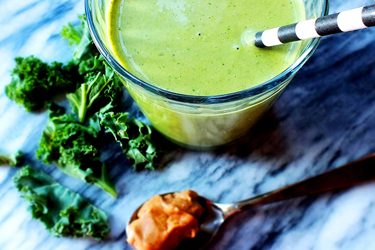 A glass filled to the brim with a green smoothie sits on a marble countertop next to a heading spoonful of peanut butter and kale leaves