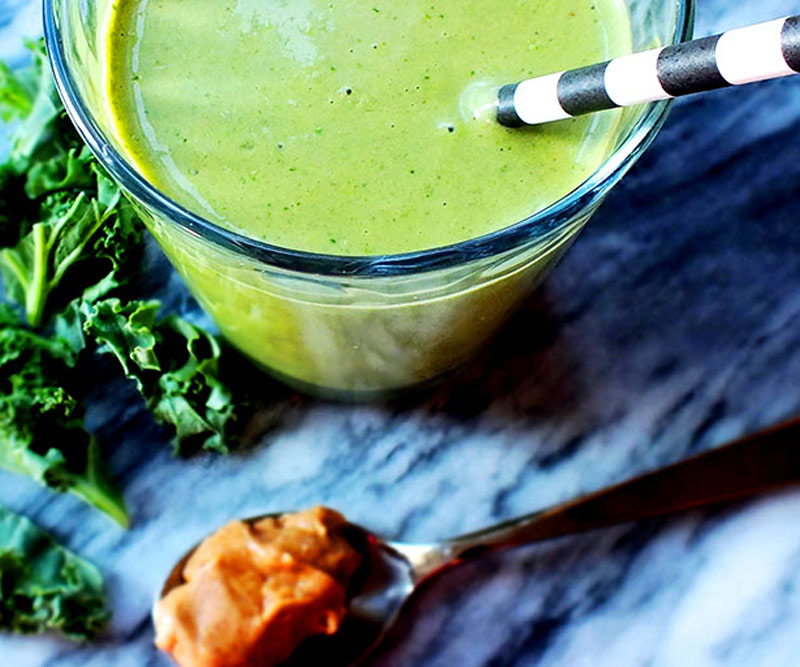 A glass filled to the brim with a green smoothie sits on a marble countertop next to a heading spoonful of peanut butter and kale leaves