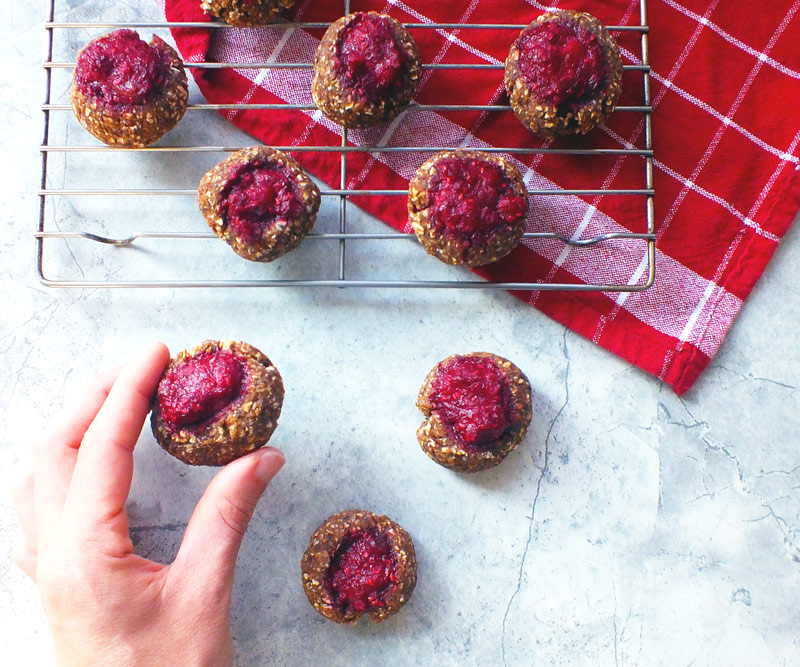 A hand reaches for a freshly made Raspberry Thumbprint Cookie resting on a wire cooling rack over a red dish towel