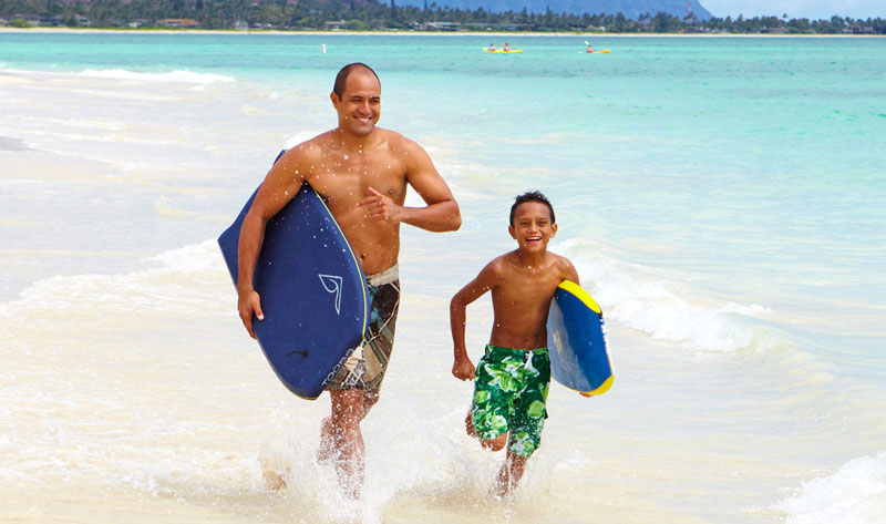 Adult male and young boy running on the beach holding boogie boards