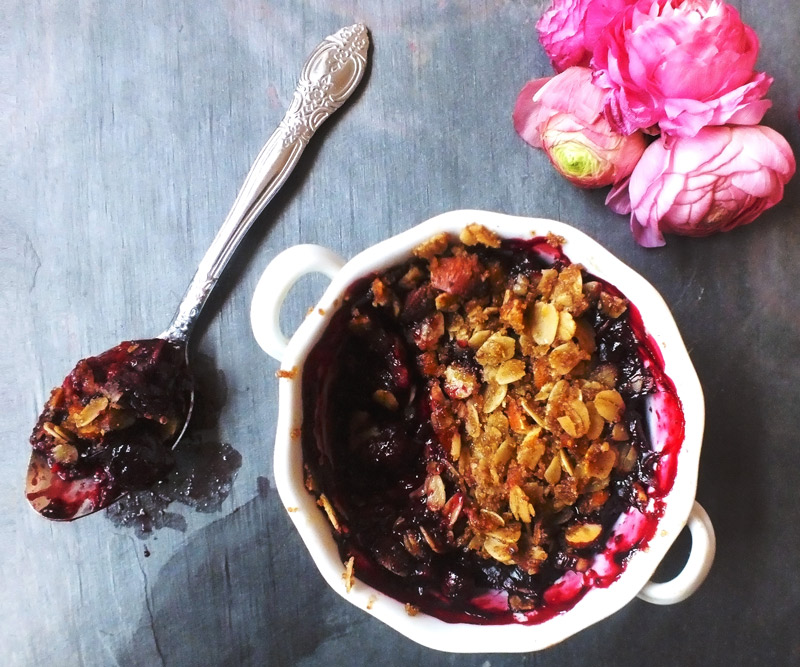 a white ramekin of Old-Fashioned Fruit Crumble with a spoonful of crumble taken out of it sits on a slate counter top that has been decorated with a bunch of pink flowers