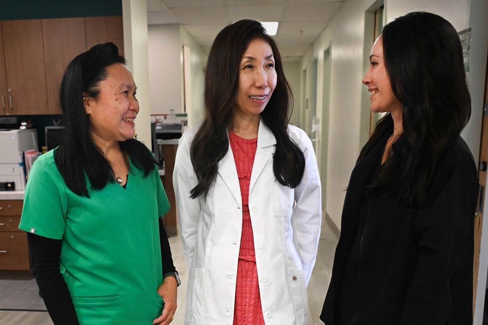 Doctor in white coat beside two colleagues, an MA in green scrubs and a pharmacist in black.