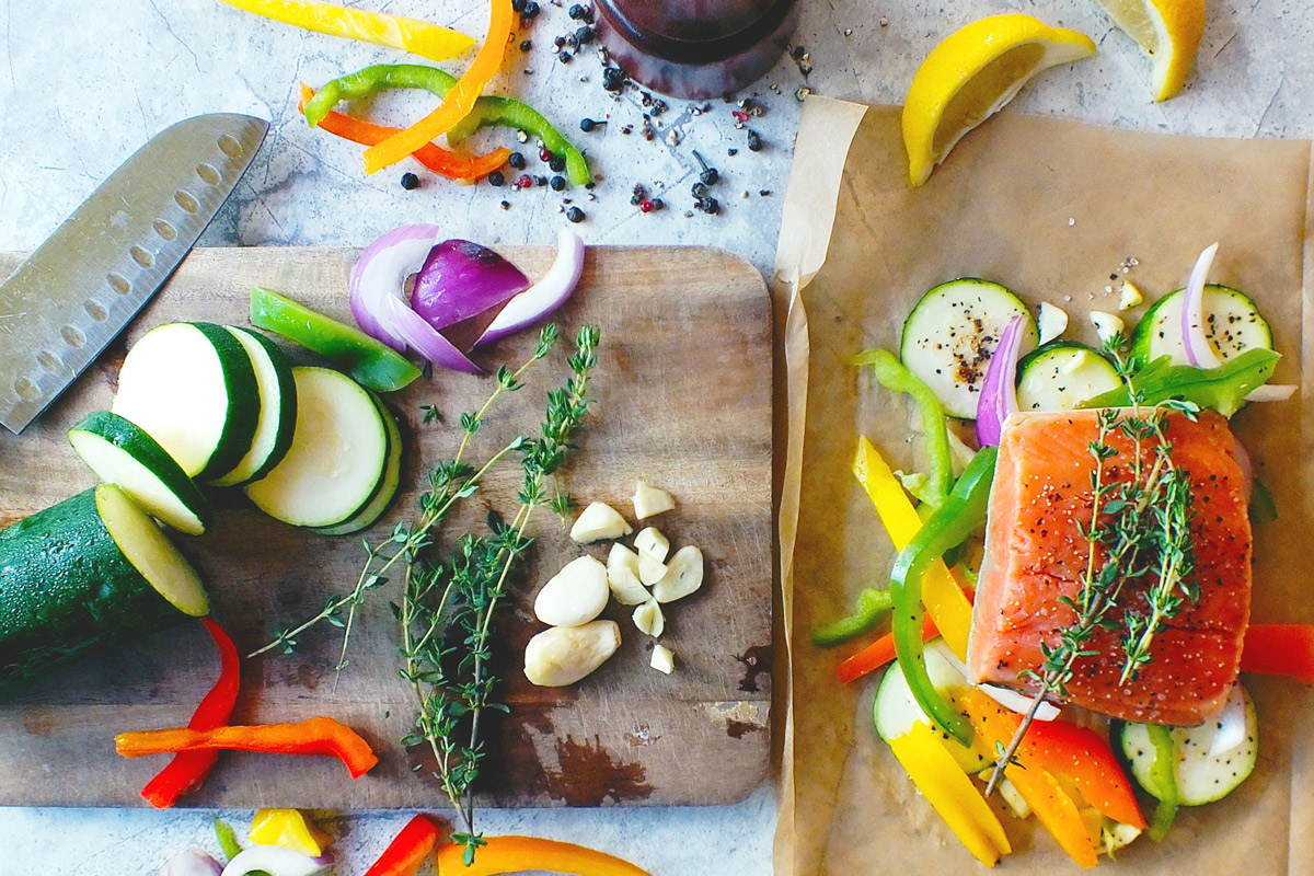 bright orange salmon arranged on a sheet of parchment paper, surrounded by sliced zucchini, bell peppers and onions next to a cutting board with more freshly sliced veggies, garlic and herbs