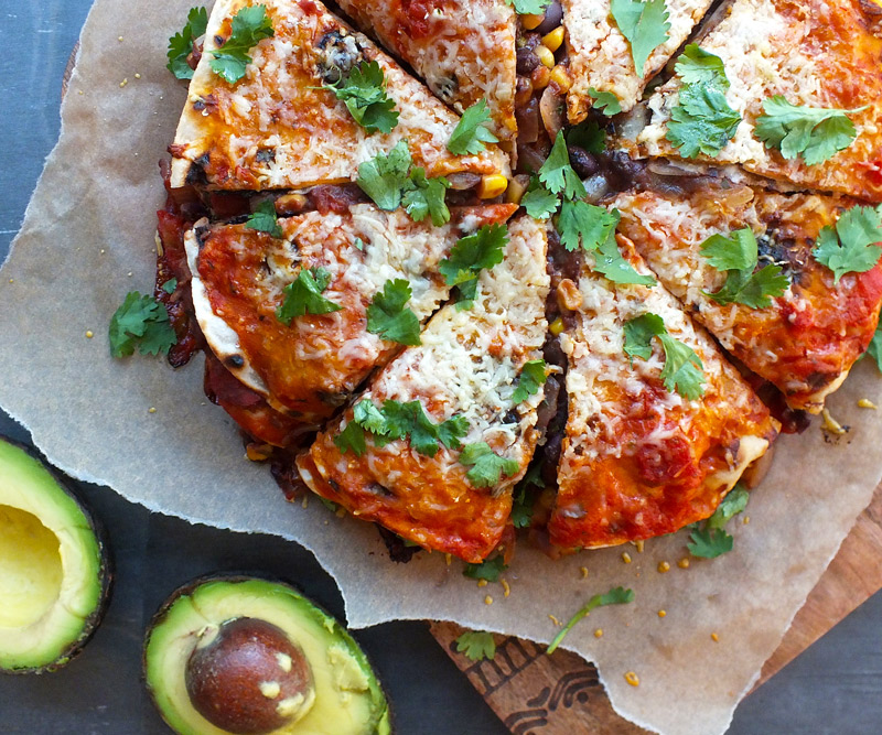 A freshly made Tex-Mex Tortilla Pie is presented on a wooden cutting board with an avocado, cut in half, next to it