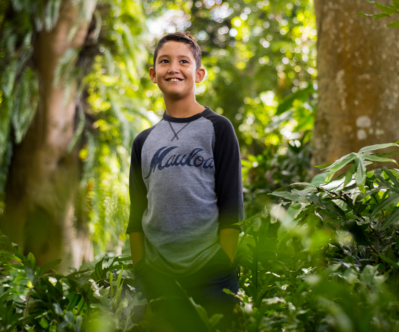 A young boy is standing in the middle of a forest wearing a baseball jersey and smiling.