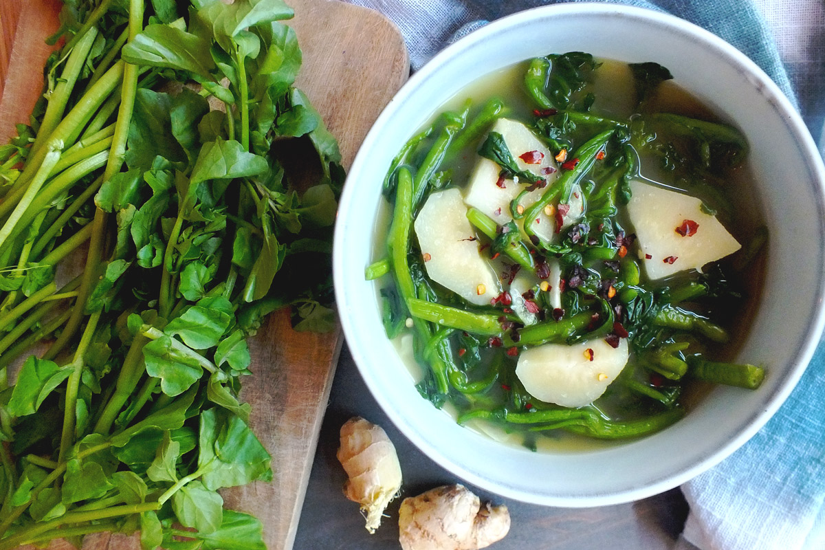 A bowl of Watercress & Ginger Soup with fresh watercress and ginger artistically surrounding the bowl