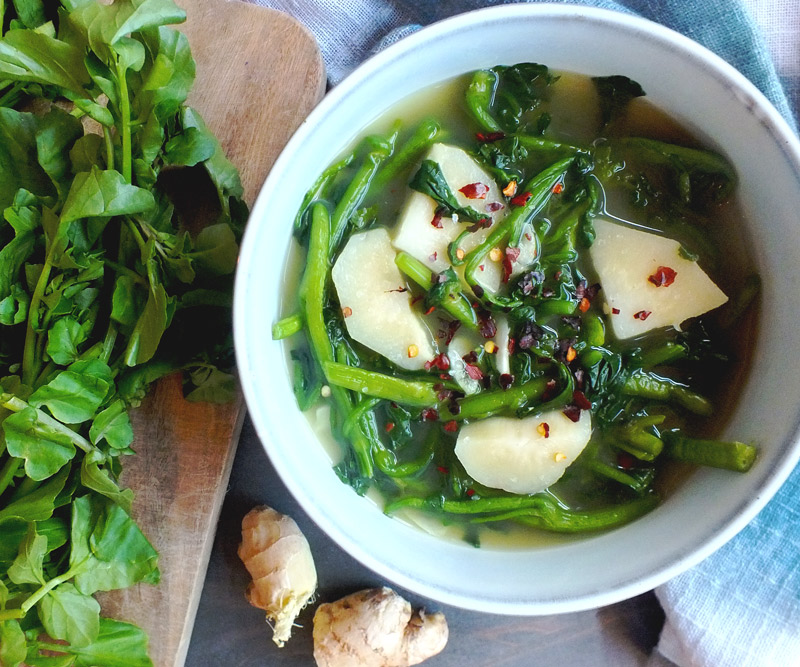 A bowl of Watercress & Ginger Soup with fresh watercress and ginger artistically surrounding the bowl