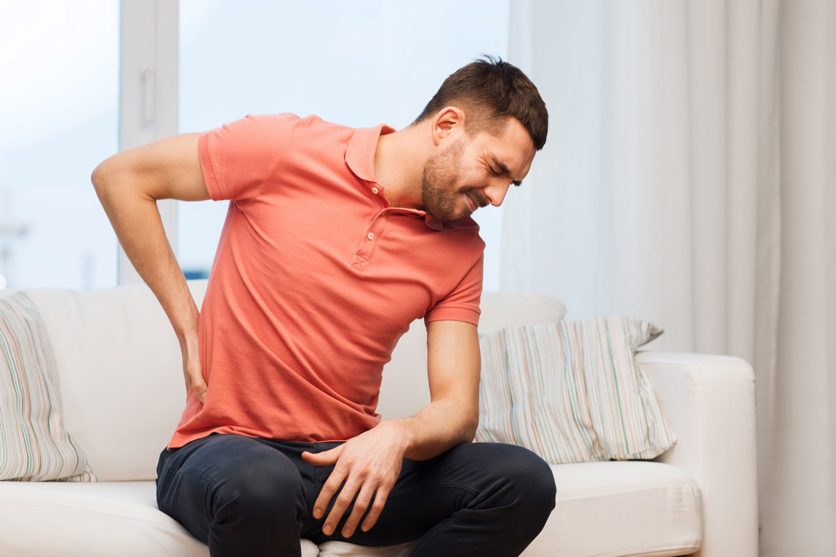 A young man sitting on a couch holding his lower back in pain