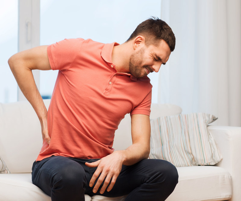 A young man sitting on a couch holding his lower back in pain