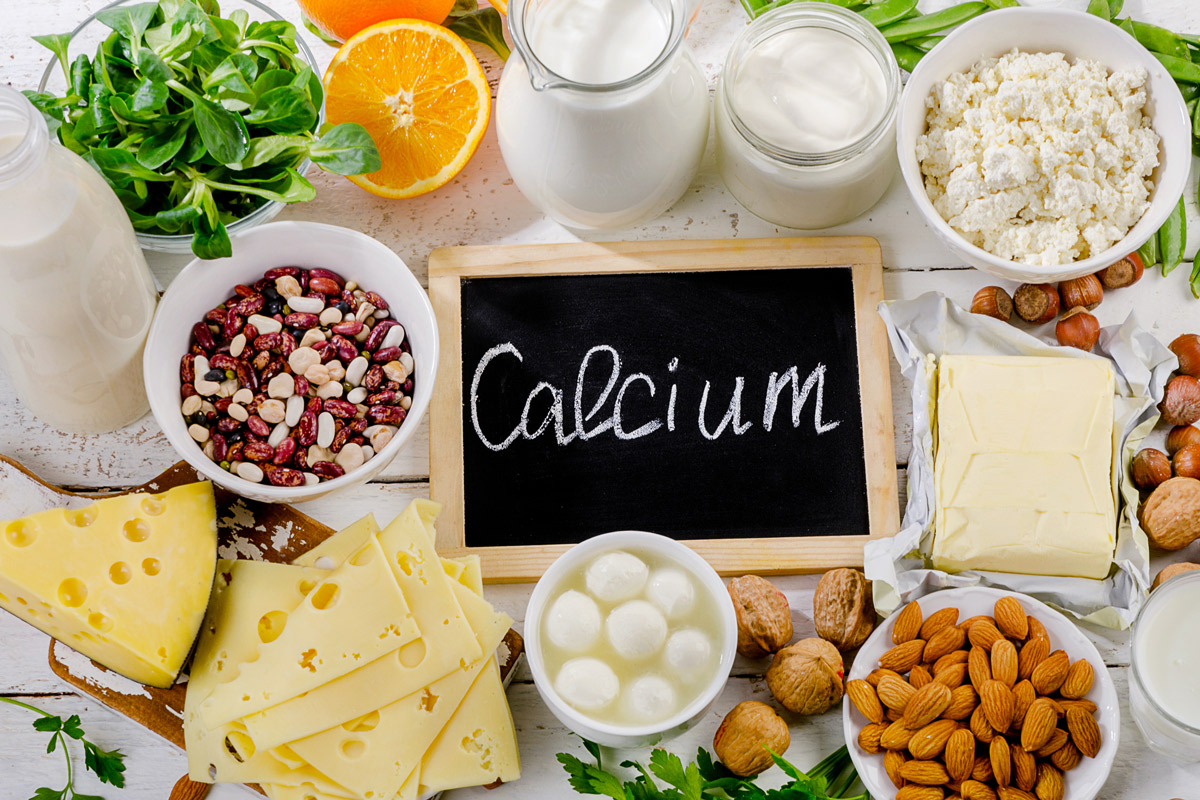 Calcium-rich foods, including milk, cheese, nuts, and leafy greens, displayed on a table around a handheld chalkboard with the word "Calcium" written on it