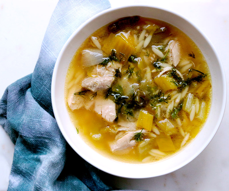 A bowl of Lemony Chicken & Orzo Soup sits on a white counter next to a blue tie-dye napkin