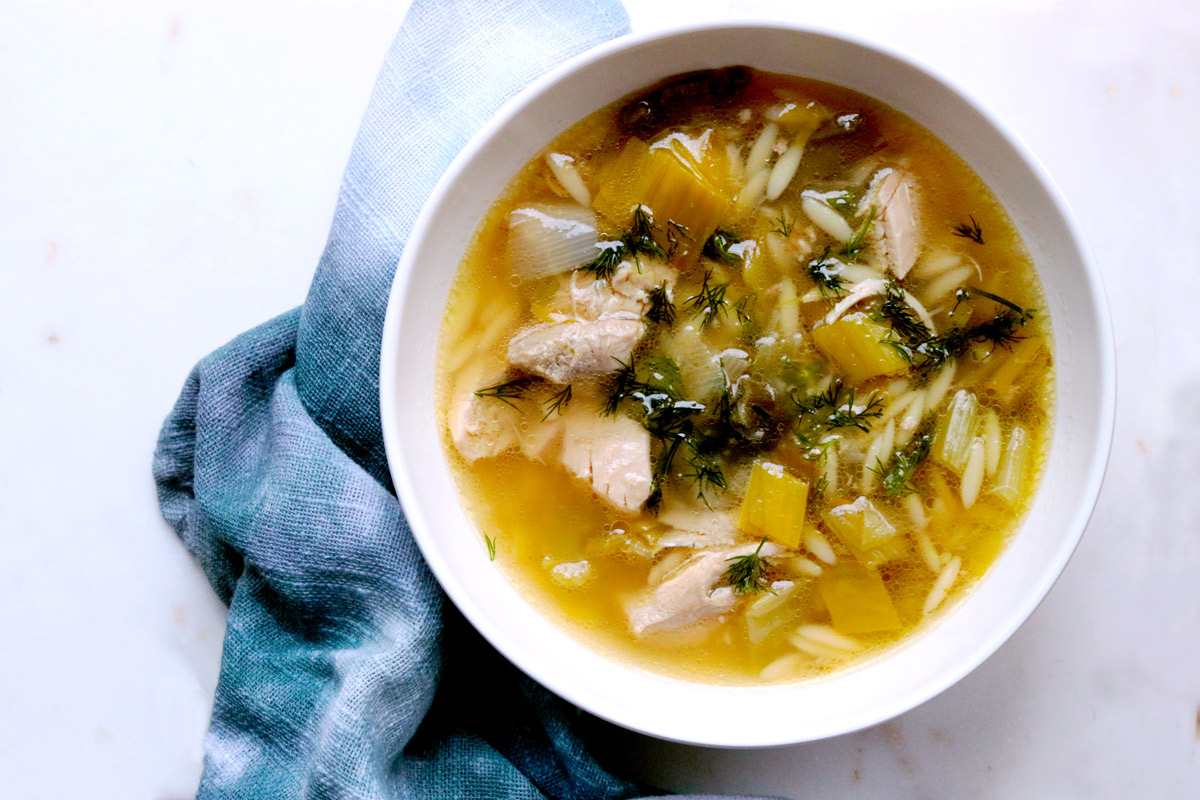 A bowl of Lemony Chicken & Orzo Soup sits on a white counter next to a blue tie-dye napkin