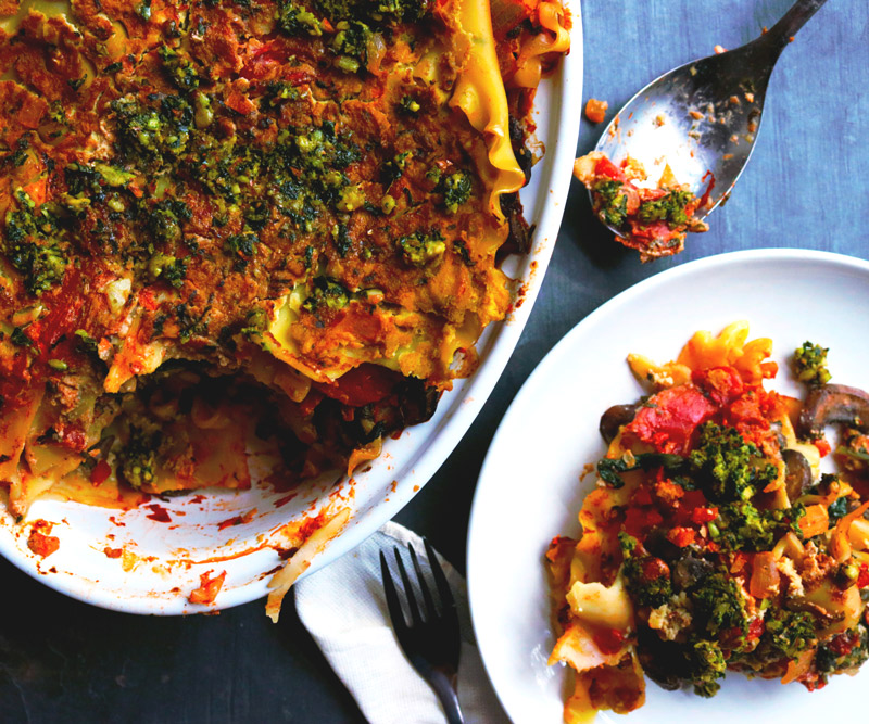 A slice of Spinach & Mushroom Lasagna sits on a plate on a counter next to a casserole dish of more lasagna with a piece taken out of it