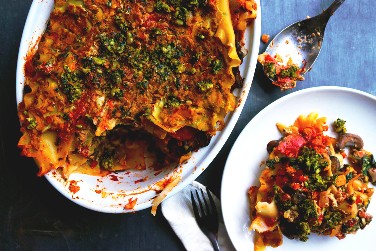 A slice of Spinach & Mushroom Lasagna sits on a plate on a counter next to a casserole dish of more lasagna with a piece taken out of it