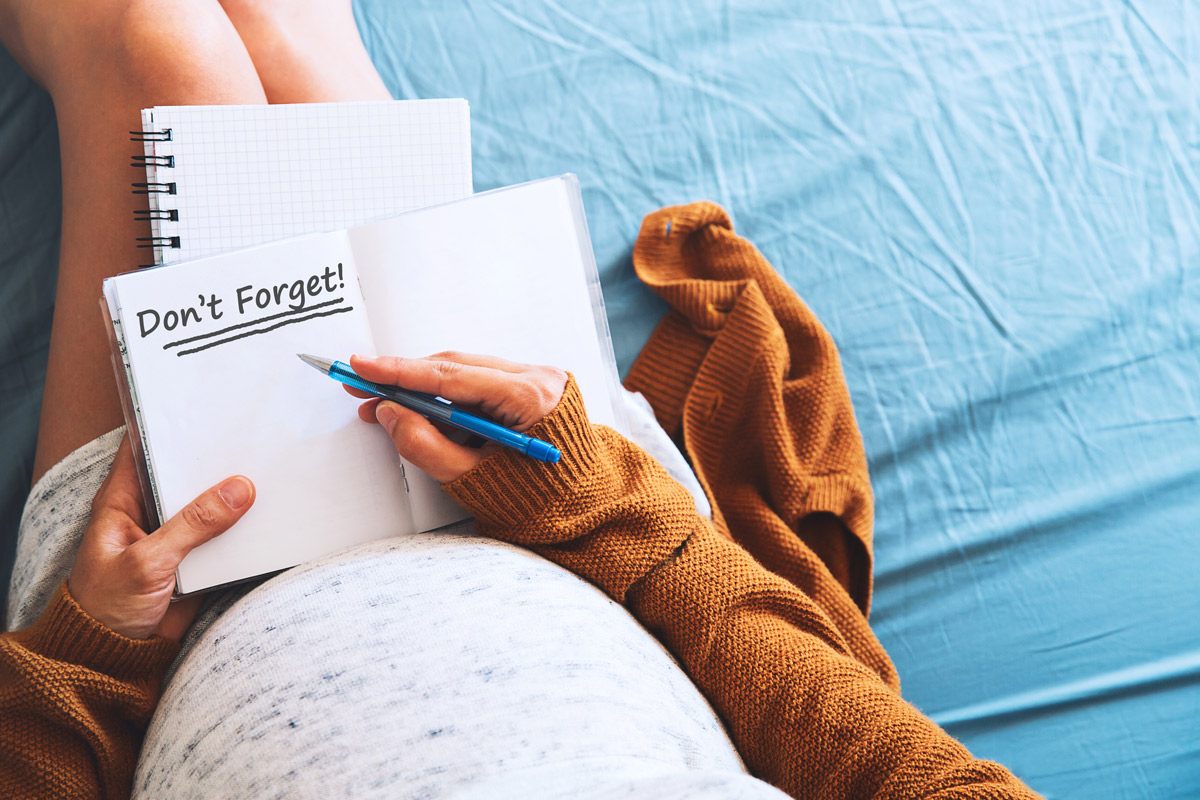 A pregnant woman sits on her bed holding a notebook with the reminder "Don't Forget!" written in bold print on the open page