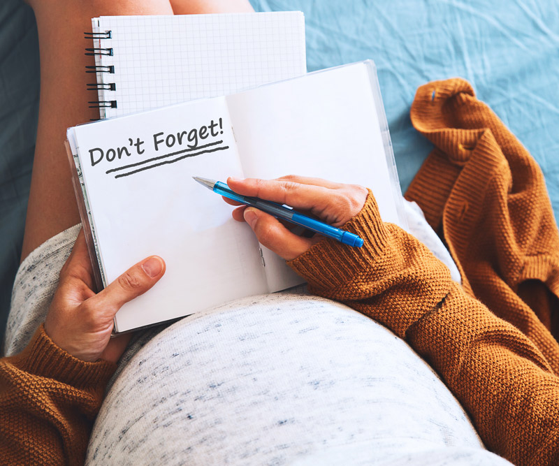 A pregnant woman sits on her bed holding a notebook with the reminder "Don't Forget!" written in bold print on the open page