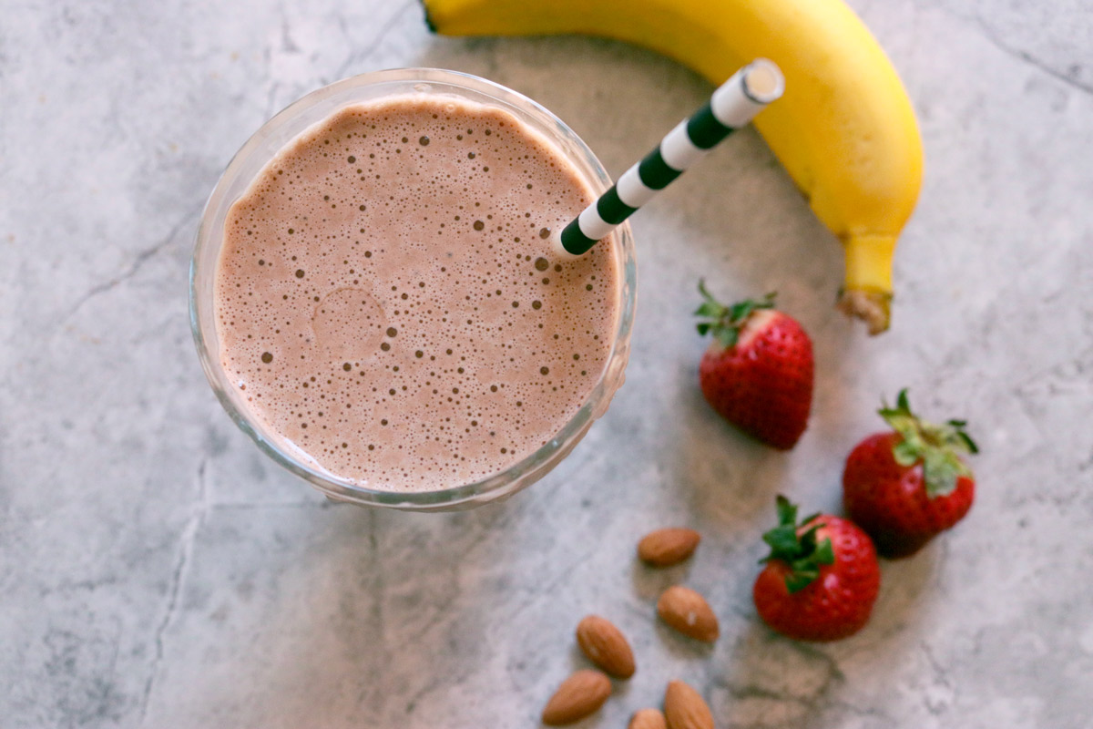 A glass full to the brim with Gail's Healthier Chocolate Protein Smoothie surrounded by a banana, fresh strawberries and almonds