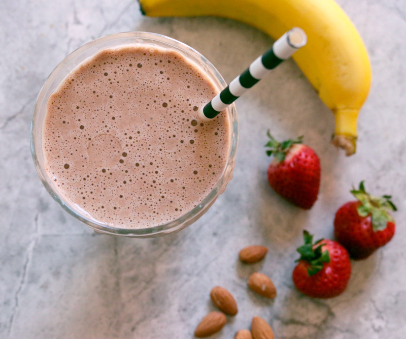 A glass full to the brim with Gail's Healthier Chocolate Protein Smoothie surrounded by a banana, fresh strawberries and almonds