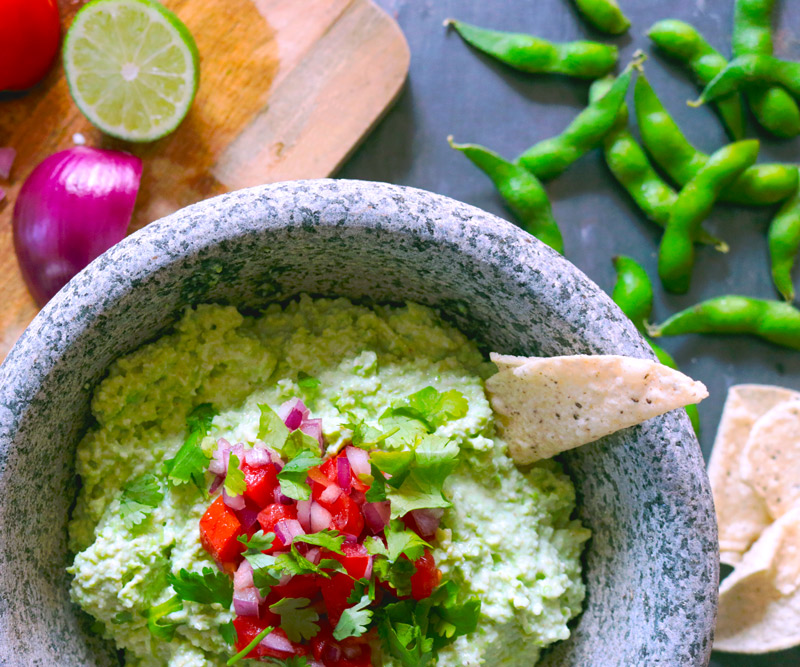 Freshly made Edamole is presented in a stone mortar topped with chopped tomatoes and red onion and a tortilla chip stick out of the dip