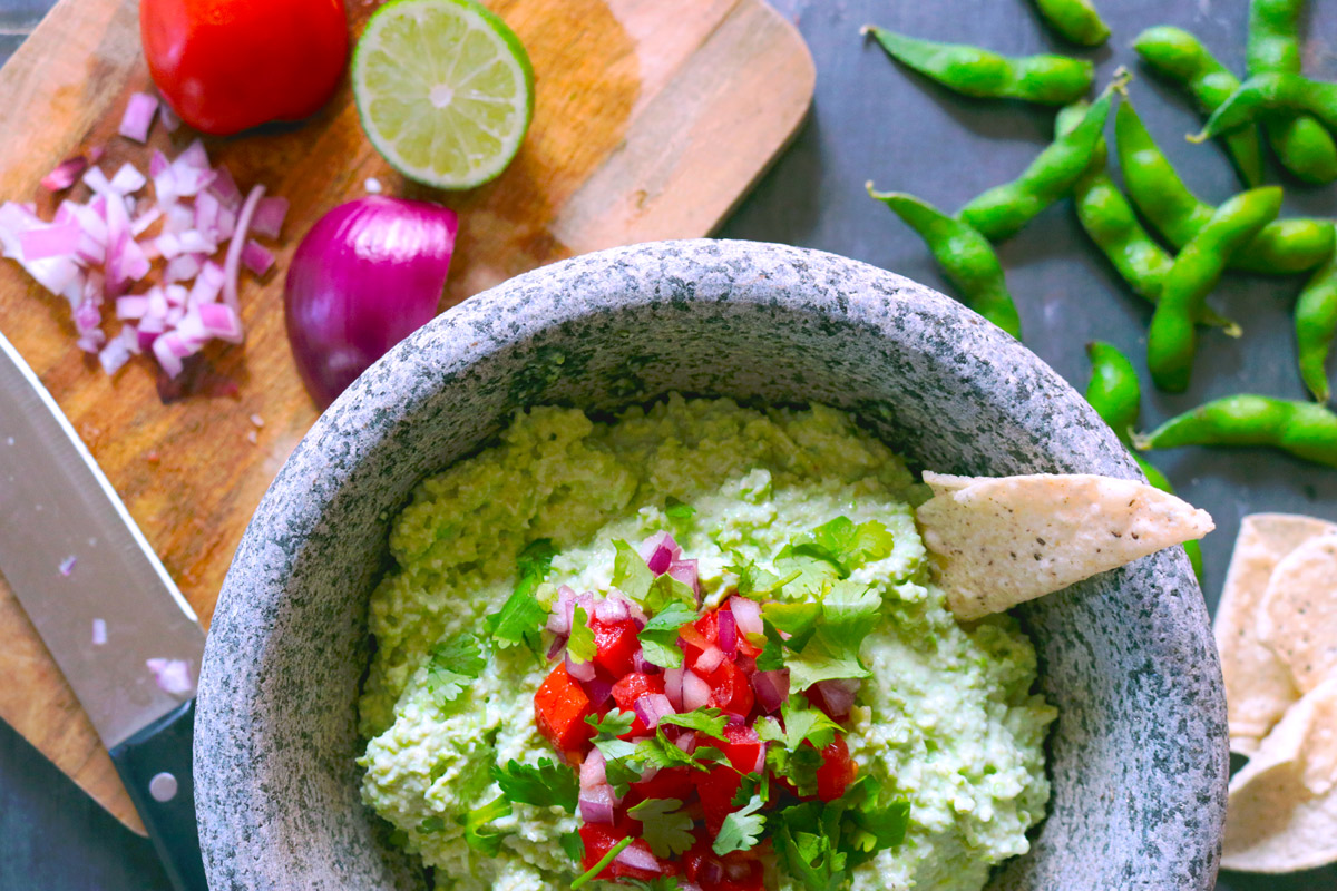 Freshly made Edamole is presented in a stone mortar topped with chopped tomatoes and red onion and a tortilla chip stick out of the dip