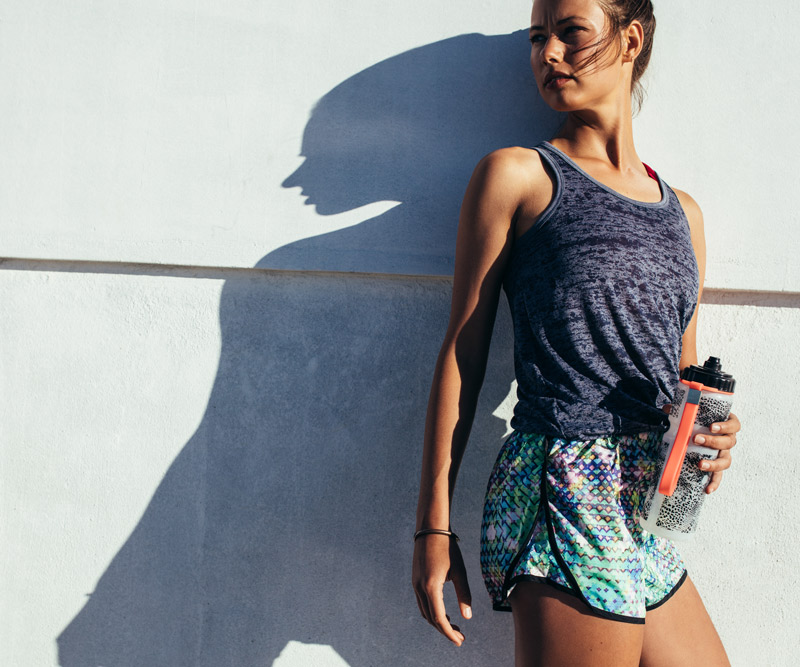 An athletic young woman stands up against a wall after a workout holding a water bottle