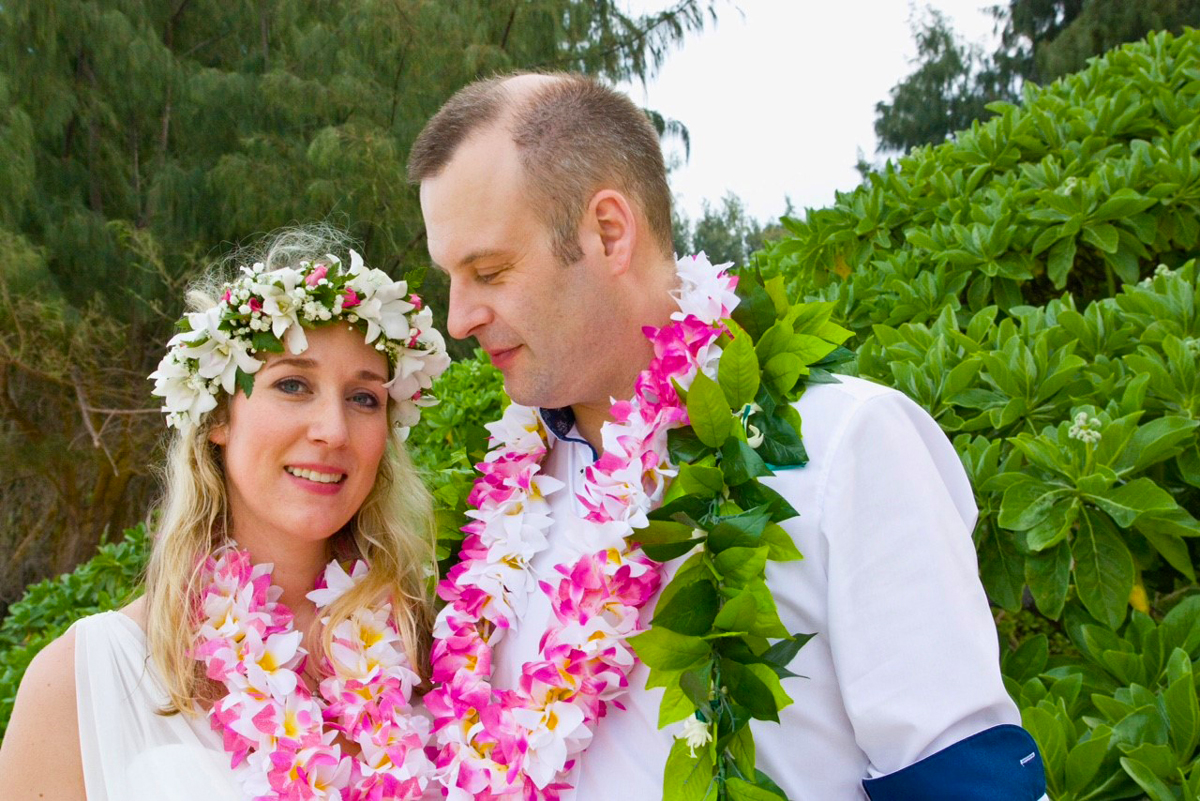 Wearing pink flower lei and dressed in white, Sinah Meier and Olli Fuchs pose for their wedding photos on the beach in Hawaii