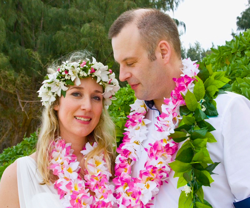 Wearing pink flower lei and dressed in white, Sinah Meier and Olli Fuchs pose for their wedding photos on the beach in Hawaii