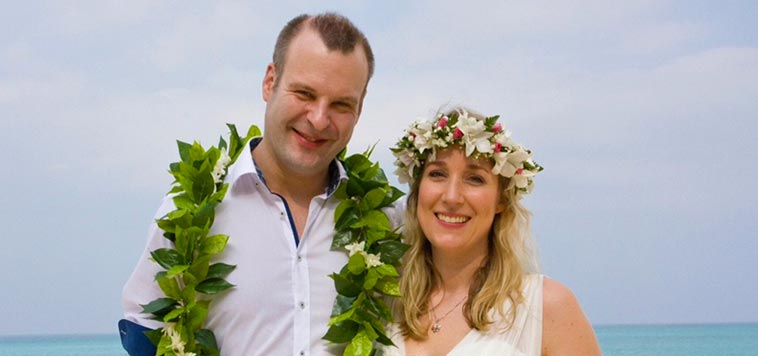 Wearing pink flower lei and dressed in white, Sinah Meier and Olli Fuchs pose for their wedding photos on the beach in Hawaii