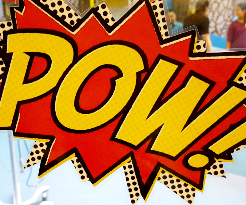 blurred silhouettes of people can be seen behind a comic book decal that says "pow" stuck to a glass window of the playroom