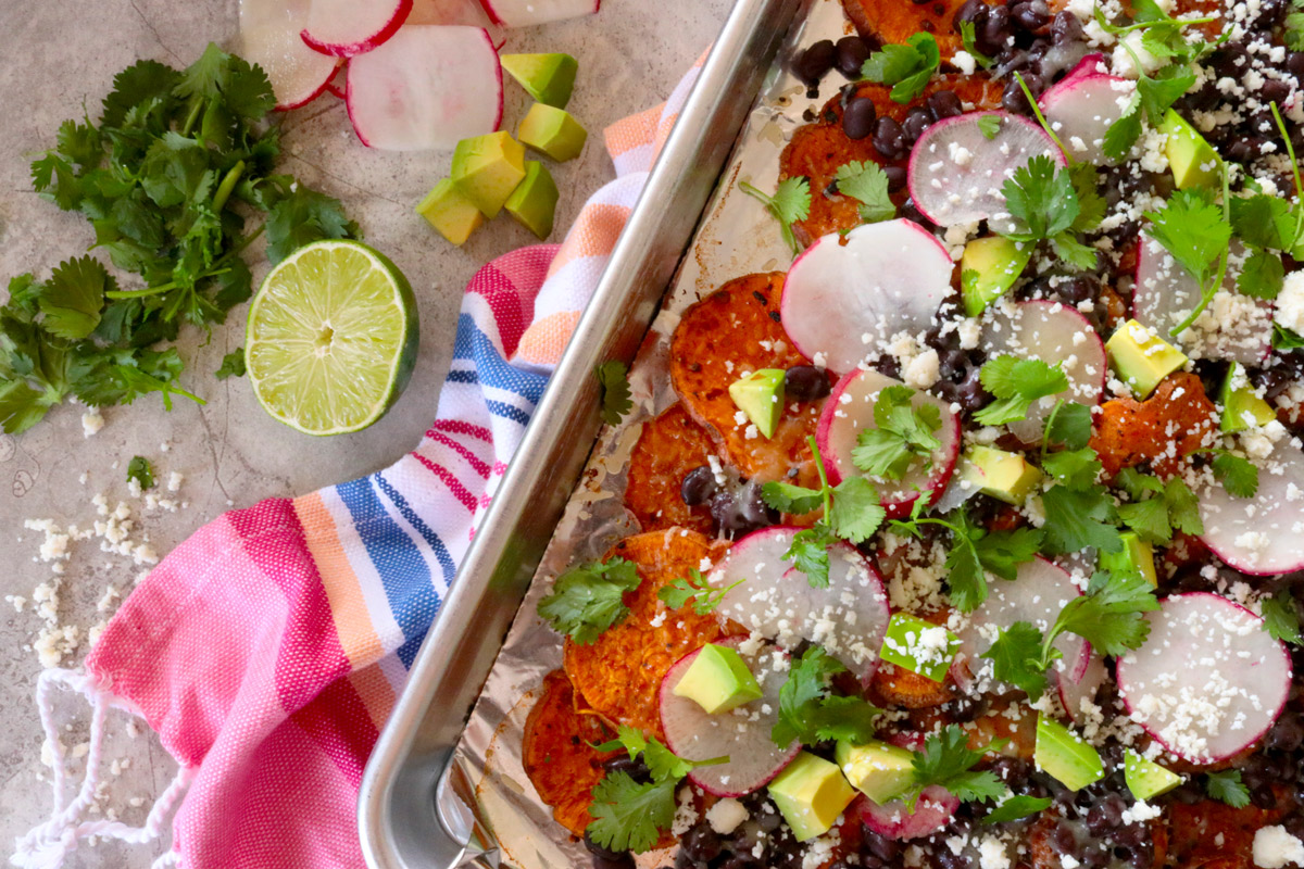 a sheet pan of Loaded Sheet Pan Sweet Potato Nachos sits on a brightly colored hand towel on a kitchen counter