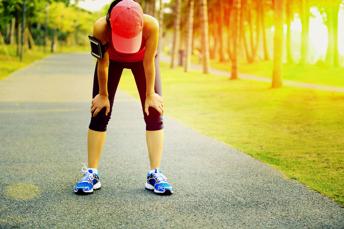 young athletic woman bent over with hands on knees as sun beats down on her back