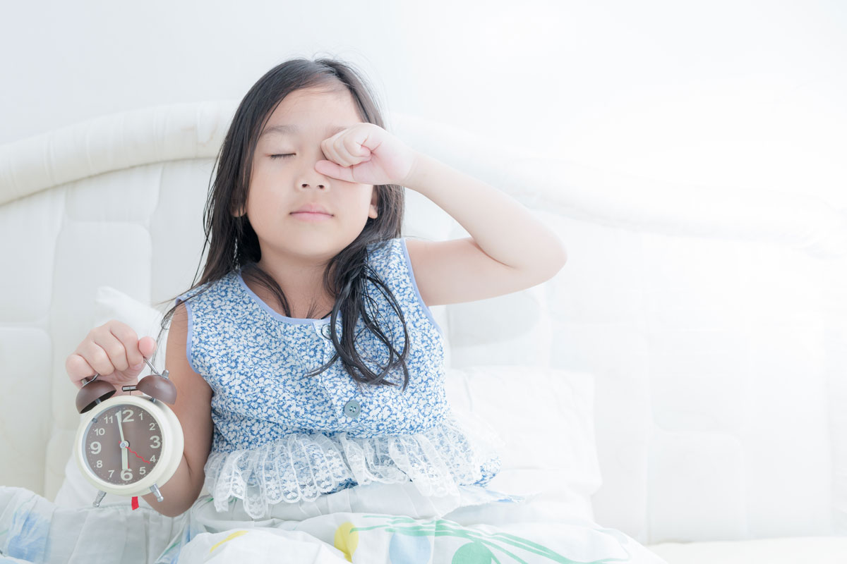 young girl sitting up in bed holding an alarm clock and rubbing sleep from her eyes