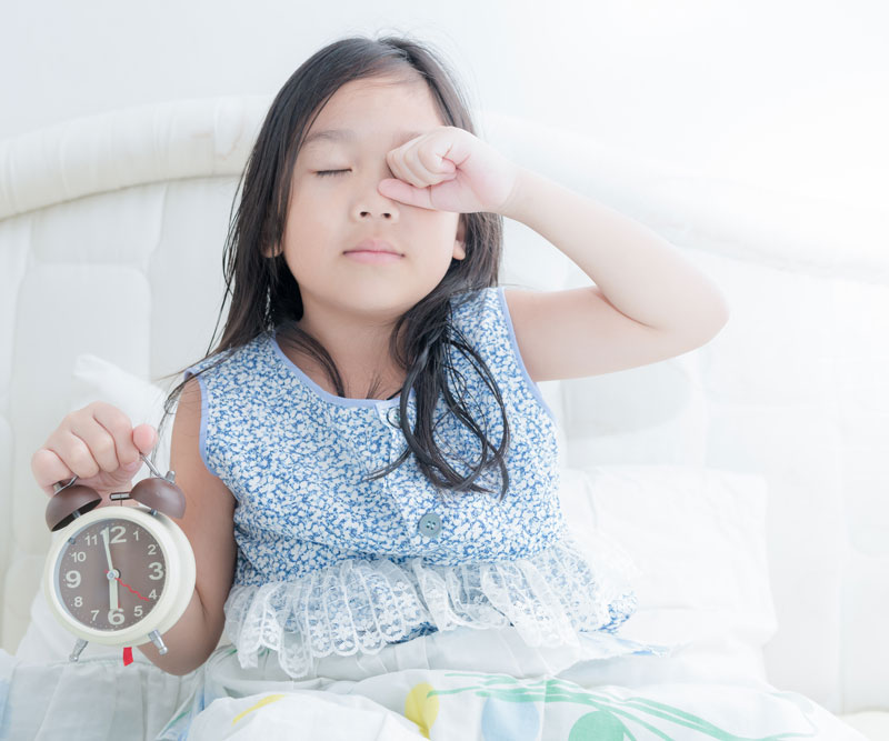 young girl sitting up in bed holding an alarm clock and rubbing sleep from her eyes