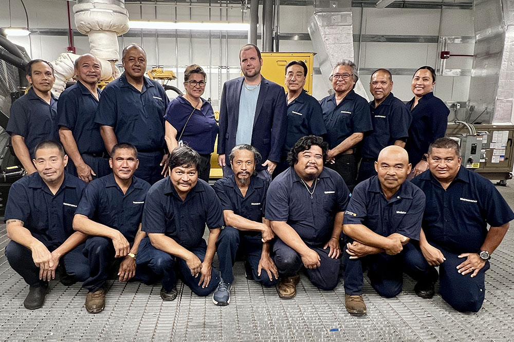 Large group with two rows of facilities staff in black polo shirts in a utility room at Wilcox Medical Center.