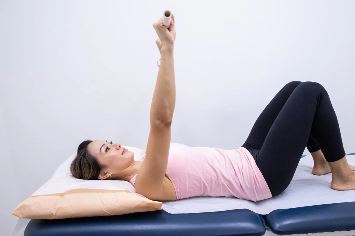 A woman in workout clothes lies on her back holding a light rod above her head.