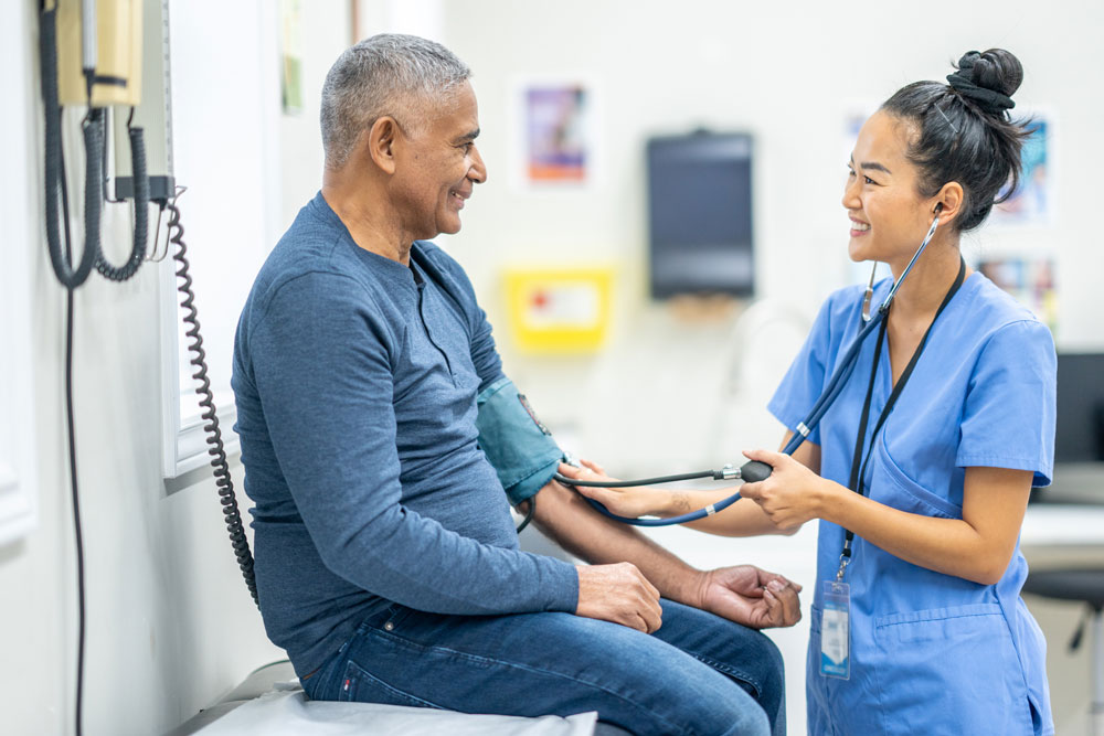 a young medical professional smiles at an older patient as she checks his blood pressure in an exam room.