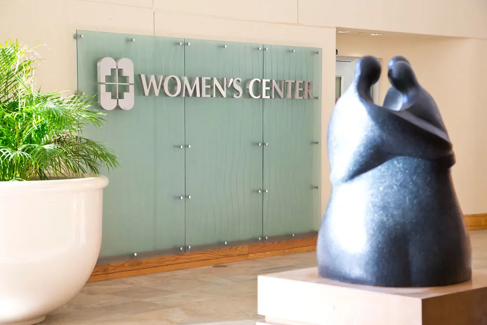 A sign for Women's Center next to a large potted plant and a stone statue of three people hugging.
